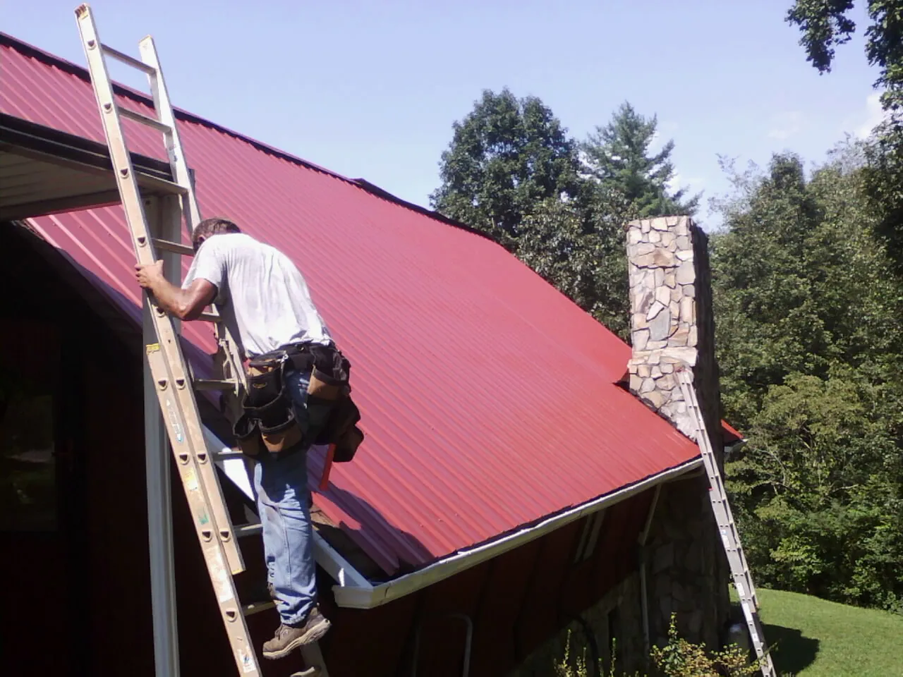 Roofer on a ladder beside a red metal roof and stone chimney — Soffit & Fascia Repair by Roofing Brothers in Armington